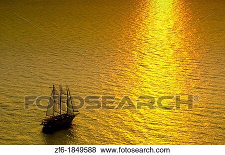 Stock Photo - Big masked sailboat sailing in sunset of beautiful Oia in Santorini Greece in Greek Islands from above on cliff into water. Fotosearch