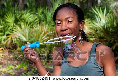 Stock Image - Black African American girl aged 18 blowing bubbles in yard outdoors laughing. Fotosearch