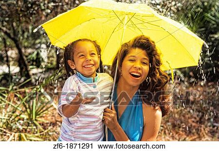 Stock Image - Black african american mother with young daughter in rain together under umbrella, laughing and wet. Fotosearch