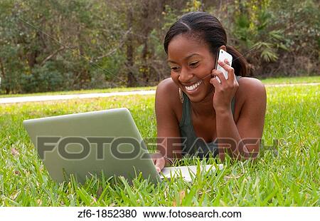 Stock Image - Black African American teen girl age 18 working on computer facebook with emailing friends cell phone in front yard on grass. Fotosearch