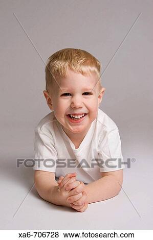 Blond haired, blue-eyed 3-yera-old boy lying on his stomach, hands clasped and smiling at the camera, wearing white t-shirt. Studio lighting, grey background. View Large Photo Image Stock Photo - Blond haired, blue-eyed 3-yera-old boy lying on his stomach, hands clasped and smiling at the camera, wearing white t-shirt. Studio lighting, grey background.. Fotosearch