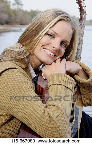 Stock Image - Blond woman holding a guitar sitting down by the lake smiling at the camera. Fotosearch