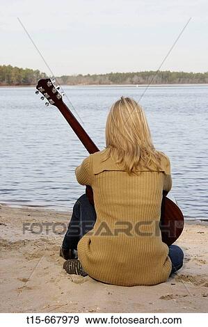 Stock Photo - Blond woman holding a guitar sitting down by the lake looking at the lake. Fotosearch
