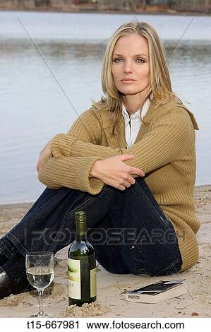 Stock Image - Blond woman in her 20's 30's sitting by a lake with wine bottle, glass and a book looking sad, tired. Fotosearch