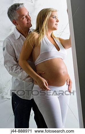 Stock Image - Blonde pregant woman wearing white standing with man behind her. Fotosearch