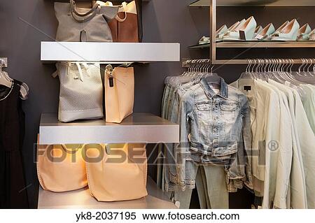 Stock Photography - Blouses, shirts and jackets hanging on rack in retail shop interior. Bags, handbags, shoulder bags on shelf.. Fotosearch