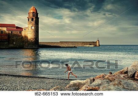 Stock Image - Boy on the beach and Notre-Dame des Anges church in background, Collioure. Roussillon-Languedoc, Pyrenees Orientales, France. Fotosearch
