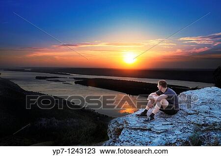 Stock Image - Boy watches sunset skies in Russian National Park sitting on a steep rock of Zhiguli mountains. Fotosearch