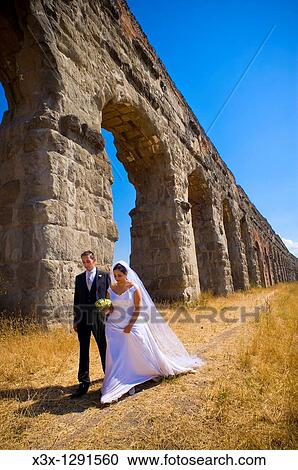Stock Image - Bride and groom walking together in under ancient aqueduct roman ruins in Rome Italy. Fotosearch