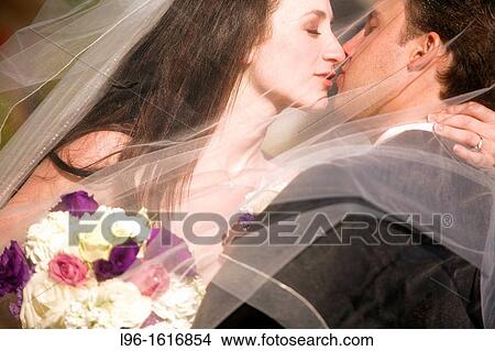 Picture - bride groom kissing embracing after wedding behind her wedding veil. Fotosearch