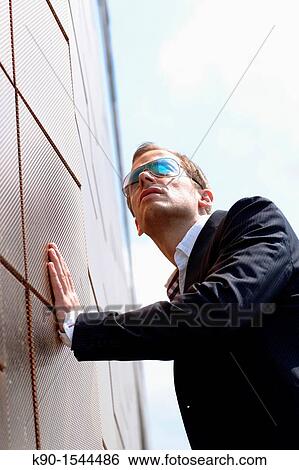 Stock Photograph - Brown haired young man with sunglasses, looking into the far distance, supporting himself by keeping one hand at a wall, Hamburg, Germany, Europe. Fotosearch