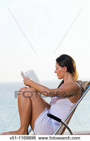 Stock Photograph - Brunette woman enjoying a book in a terrace with views at the sea. Fotosearch