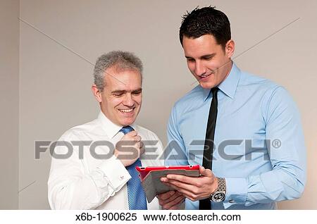 Stock Photography - Business colleagues, one young and one older, discuss work using tablet computer. Fotosearch