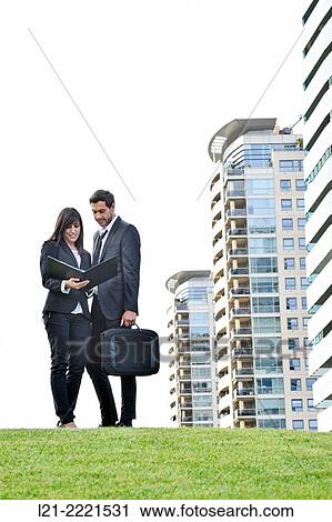 Stock Image - Businessman and businesswoman look over paperwork while standing outside.. Fotosearch