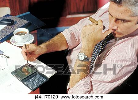 Businessman sitting at a desk with cigar and electronic organizer View Large Photo Image Stock Photo - Businessman sitting at a desk with cigar and electronic organizer. Fotosearch
