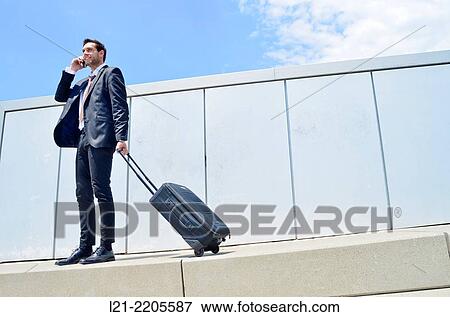 Businessman, standing, talking with a mobile while holding a suitcase on wheels outdoors. View Large Photo Image Stock Photo - Businessman, standing, talking with a mobile while holding a suitcase on wheels outdoors.. Fotosearch