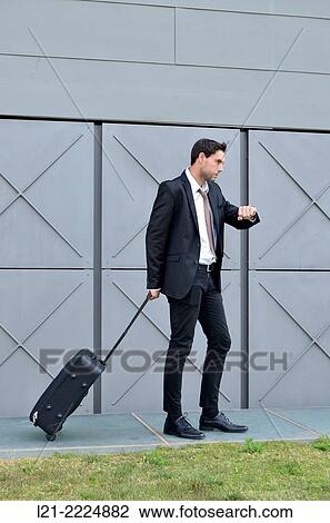 Stock Image - Businessman with wheeled suitcase looking at wrist watch.. Fotosearch