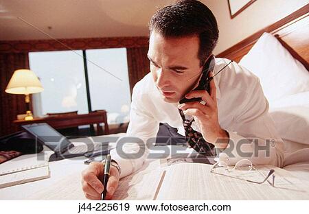Stock Photo - Businessman working on the bedon the bed and talking on cell phone in a hotel room. Fotosearch