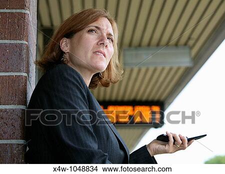 Businesswoman standing on platform hold her cell phone with train timetable in background awaiting the arrival of her commuter train on her way to work View Large Photo Image Picture - Businesswoman standing on platform hold her cell phone with train timetable in background awaiting the arrival of her commuter train on her way to work. Fotosearch