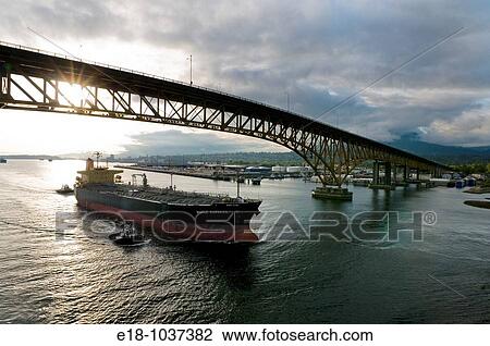 Stock Image - Canada, BC, Vancouver Freighter is escorted under the Second Narrows bridge on the Burrard Inlet by harbour tugs. Fotosearch