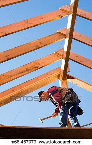 Stock Photo - Carpenter hammering trimmer to center post while standing below the framing of the roof rafters at a construction site.. Fotosearch
