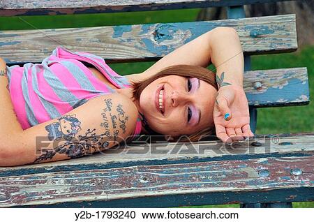 Caucasian 24 year old brown haired tattoed female model wearing pink and gray sweater lying on park bench View Large Photo Image Stock Image - Caucasian 24 year old brown haired tattoed female model wearing pink and gray sweater lying on park bench. Fotosearch