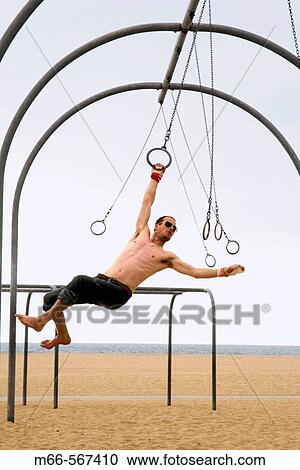 Stock Image - Caucasian man on the rings set, swinging from one ring to the next. Santa Monica Beach, Los Angeles, California.. Fotosearch