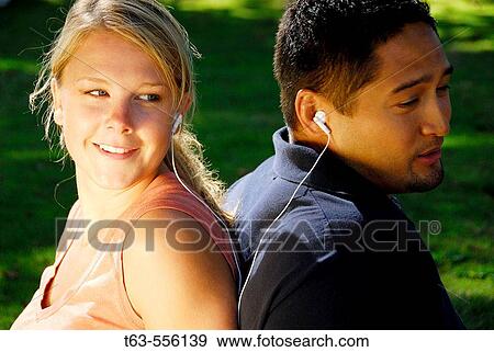 Stock Photo - caucasion female and ethnic male sitting back to back sharing music earphones. Fotosearch