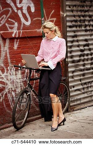 Stock Image - Caucassian businesswoman working on laptop leaning on bicycle against a graffiti wall. Fotosearch