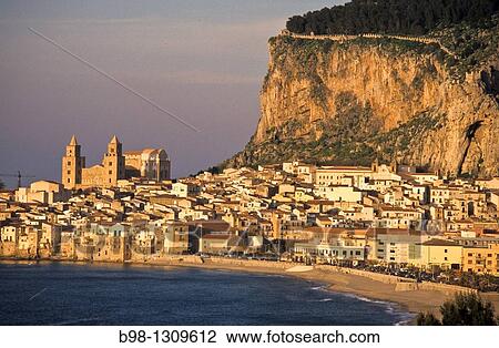 Stock Image - Cefalu, Sicily, Italy. Fishing harbor of the medieval town in late afternoon light. Boats tied to dock. Village and giant rock? la rocca? in background, province of Palermo.. Fotosearch