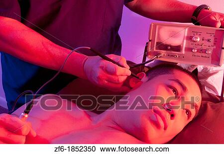 Stock Image - Chinese Acupuncture Doctor giving male patient acupuncture in ear with electronic vibrating stimulation with Stim Flex 400 machine. Fotosearch