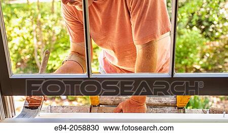 Stock Image - Close in detail of a 41 year old man painting the window trim of a house facing inside.. Fotosearch
