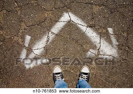Stock Image - Close-up of a man's feet standing next to two arrows painted on a driveway. Fotosearch