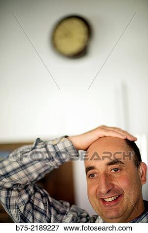Stock Photo - Close-up of the face of a 40-year-old Caucasian man, smiling and with positive attitude, with one hand on his head and wearing plaid shirt and a clock in the background.. Fotosearch