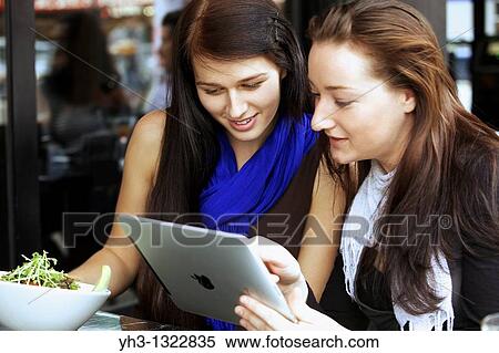 Stock Photography - Close up of two beautiful young woman reading from an iPad at a restaurant or cafe. Fotosearch