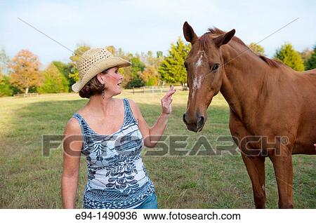 Stock Photograph - Close up portrait of a 52 year old blonde woman wearing a tank top and a straw hat interacting with a horse in a pasture.. Fotosearch