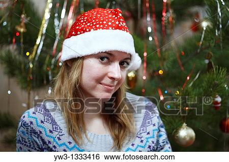 Stock Photograph - closeup of pretty girl in christmas hat against decorated xmas tree. Fotosearch