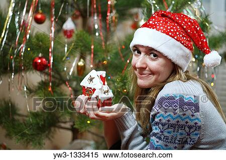 Stock Photography - closeup of pretty girl in christmas hat against decorated xmas tree with red little haus in hands. Fotosearch