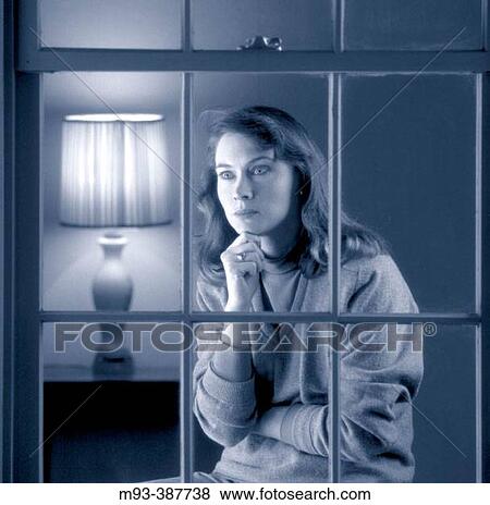 Stock Photo - Concerned or contemplative looking 30-ish woman staring out window. From outside perspective; traditional table lamp visible in background.. Fotosearch