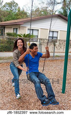 Stock Photo - Couple multi ethnics dating in park on swing with fun and Asian Hispanic ethnic laughter near home in playground. Fotosearch