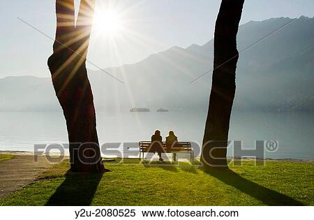 Couple sitting on a bench on the lakefront maggiore with brissago islands and mountain with sunbeam on a misty lake in ascona ticino switzerland. View Large Photo Image Stock Photography - Couple sitting on a bench on the lakefront maggiore with brissago islands and mountain with sunbeam on a misty lake in ascona ticino switzerland.. Fotosearch