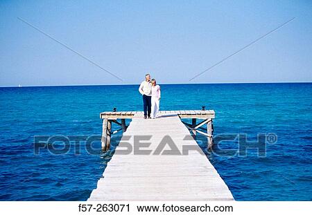 Stock Image - Couple sitting walking in a wooden footbridge over the sea. Fotosearch