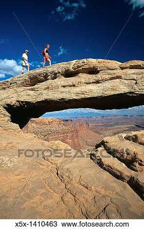 Stock Image - Couple Walking across Mesa Arch, Canyonlands National Pak, Utah, USA. Fotosearch