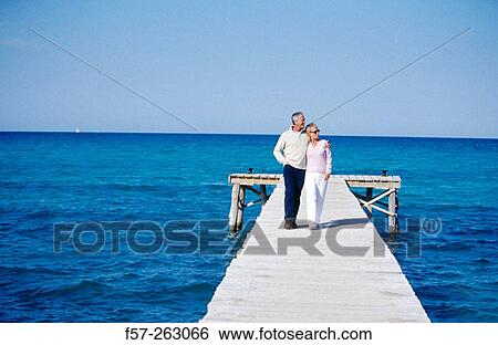 Stock Photograph - Couple walking in a wooden footbridge over the sea. Fotosearch