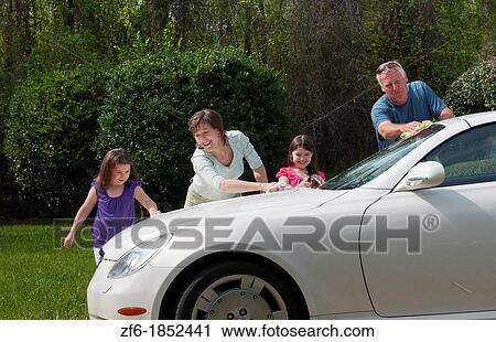 Stock Image - Dad and mom and daughters family washing car at home in driveway together with hose and having fun. Fotosearch
