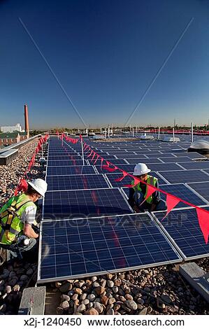 Stock Image - Denver, Colorado - Workers install solar photovoltaic panels on the roof of Harrington Elementary School The worker inside the red-flagged zone wears a fall protection harness. Fotosearch