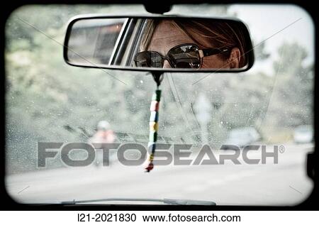 Stock Image - Detail of a woman wearing sunglasses in the rearview mirror of a car. Blurry vision of the road and vehicles through the windshield. Barcelona province, Catalonia, Spain.. Fotosearch