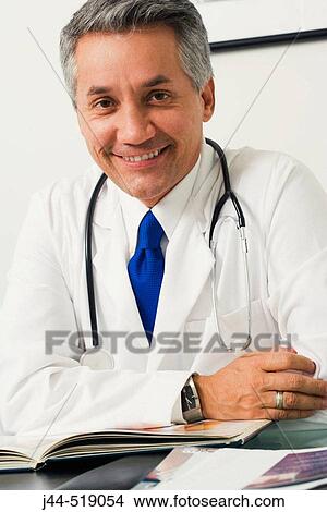 Doctor sitting on desk smiling with stethoscope on neck View Large Photo Image Picture - Doctor sitting on desk smiling with stethoscope on neck. Fotosearch