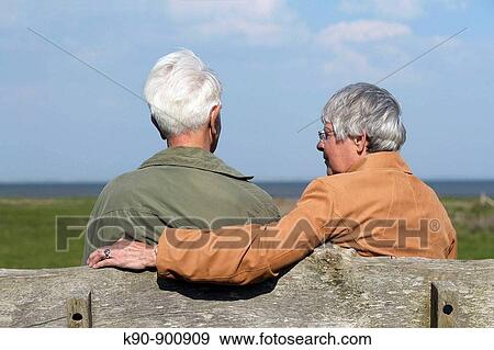 Stock Photo - Elderly couple sitting on a bench and seen from backside while the woman is looking at the man with bright blue sky in the background, Island of Amrum, Northfrisian, Schleswig-Holstein, Northern Germany, Europe. Fotosearch
