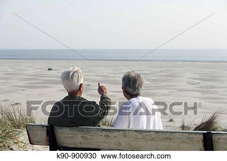 Stock Image - Elderly couple sitting on a bench, seen from backside, looking over the wide beach with the sea in the background and the senior man pointing with his finger, Island of Amrum, Northfrisian, Schleswig_Holstein, Northern Germany, Europe. Fotosearch
